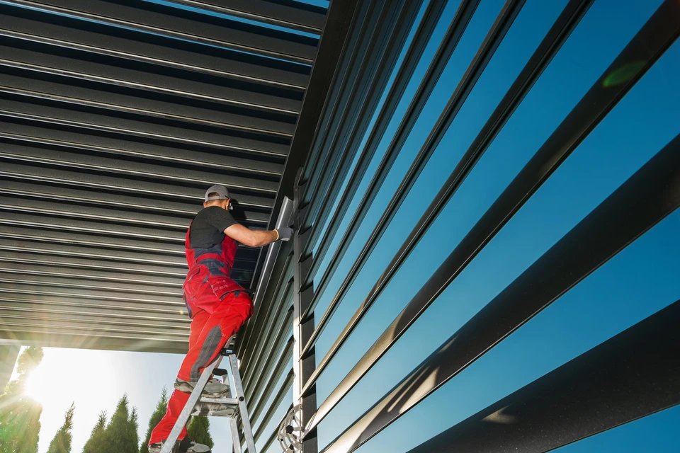 Ouvrier en combinaison rouge installant des volets roulants extérieurs sur échelle, travaux de menuiserie aluminium sur façade de bâtiment moderne sous ciel bleu