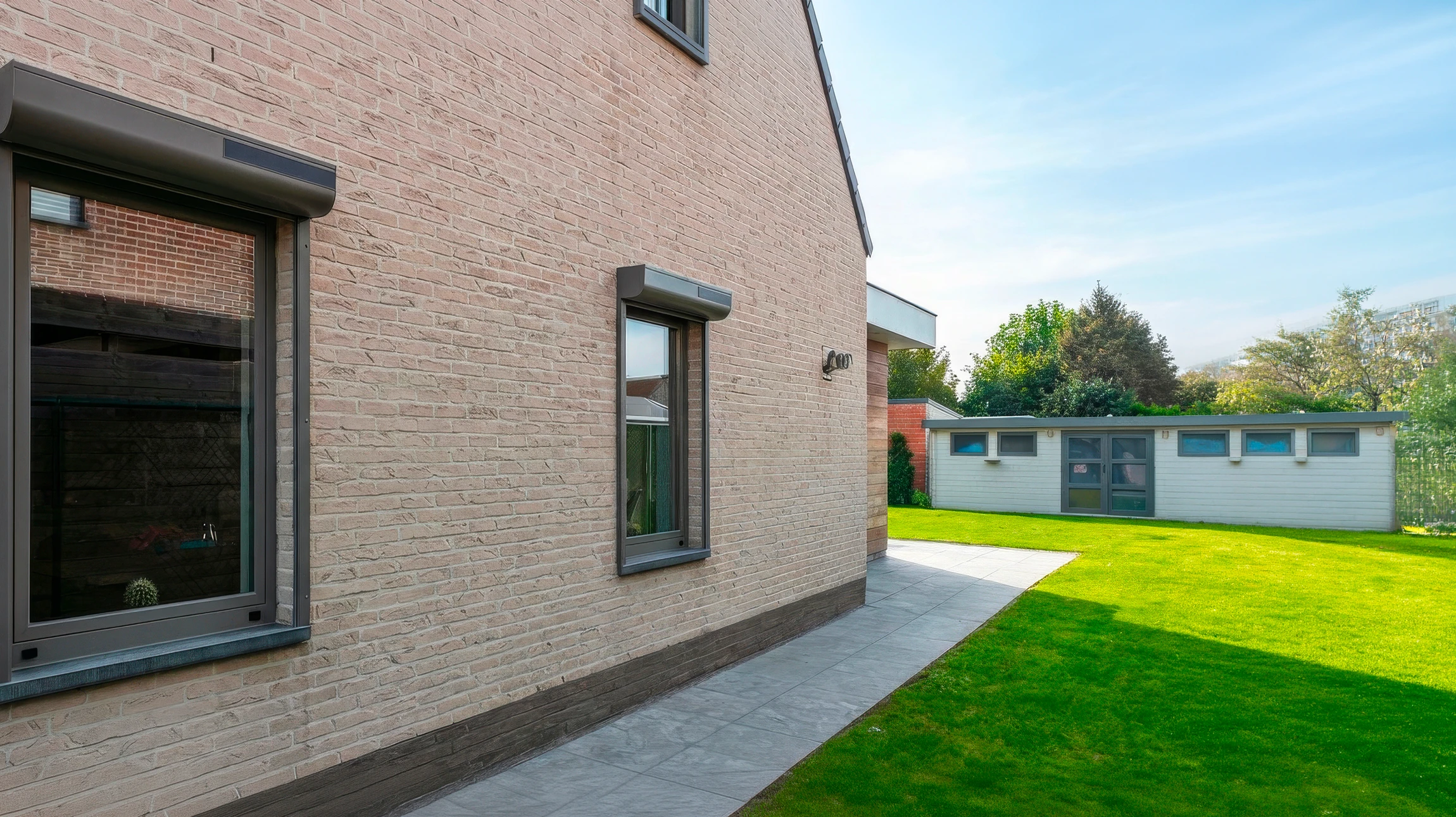 Façade latérale d'une maison contemporaine en briques beiges, avec deux fenêtres à cadres anthracite équipées de caissons de volets roulants intégrés