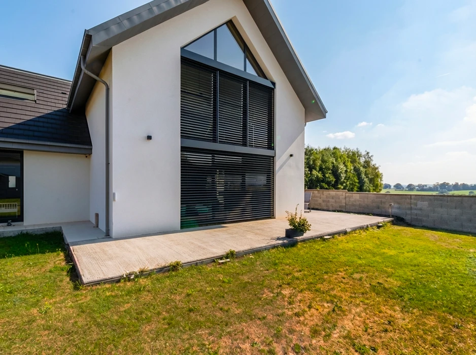 Façade arrière d'une maison contemporaine à toit en bâtière, enduit blanc, avec grande baie vitrée double hauteur sous pignon, équipée de stores vénitiens extérieurs à lames orientables en aluminium anthracite