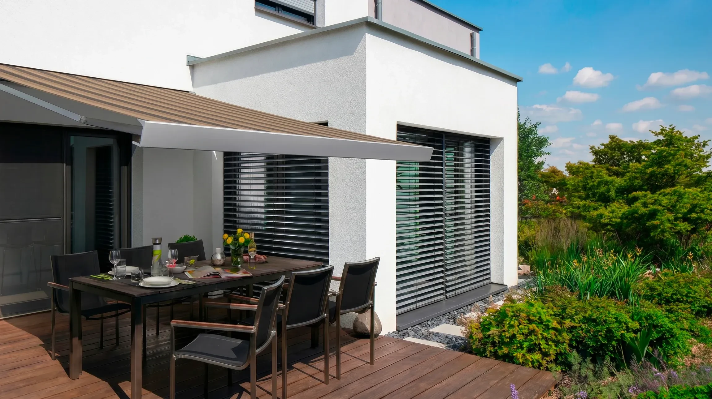 Terrasse extérieure moderne d'une maison contemporaine équipée d'un store screen enroulable de couleur gris anthracite. La toile technique microperforée est partiellement déroulée, offrant une occultation partielle tout en laissant passer la lumière. La structure de guidage est en aluminium anthracite, montée en applique sur la façade blanche de la maison. Un mobilier de jardin en bois ou aluminium et un plan d'eau (piscine ou plan d'eau paysager) sont visibles en arrière-plan, illustrant un environnement résidentiel serein et protégé des apports solaires excessifs.