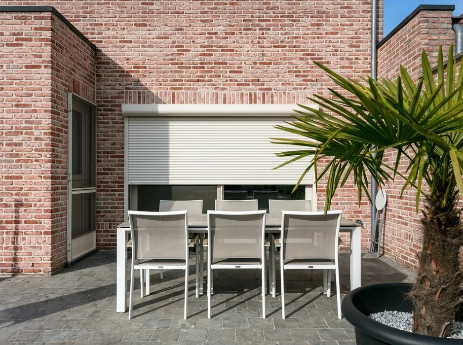 Façade de maison en briques rouges avec volet roulant aluminium couleur taupe partiellement ouvert, révélant une baie vitrée coulissante triple vantaux avec rideaux voilages blancs